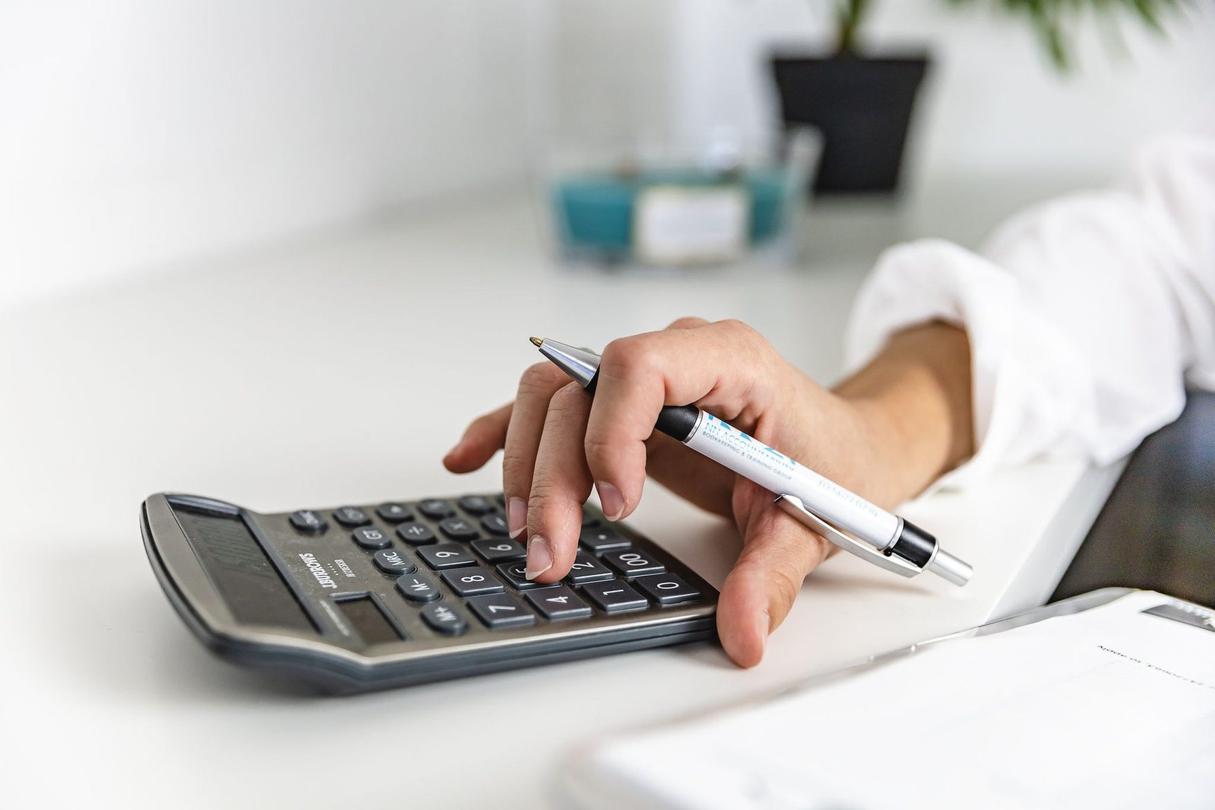 A hand working a calculator with a pen, beside a notepad