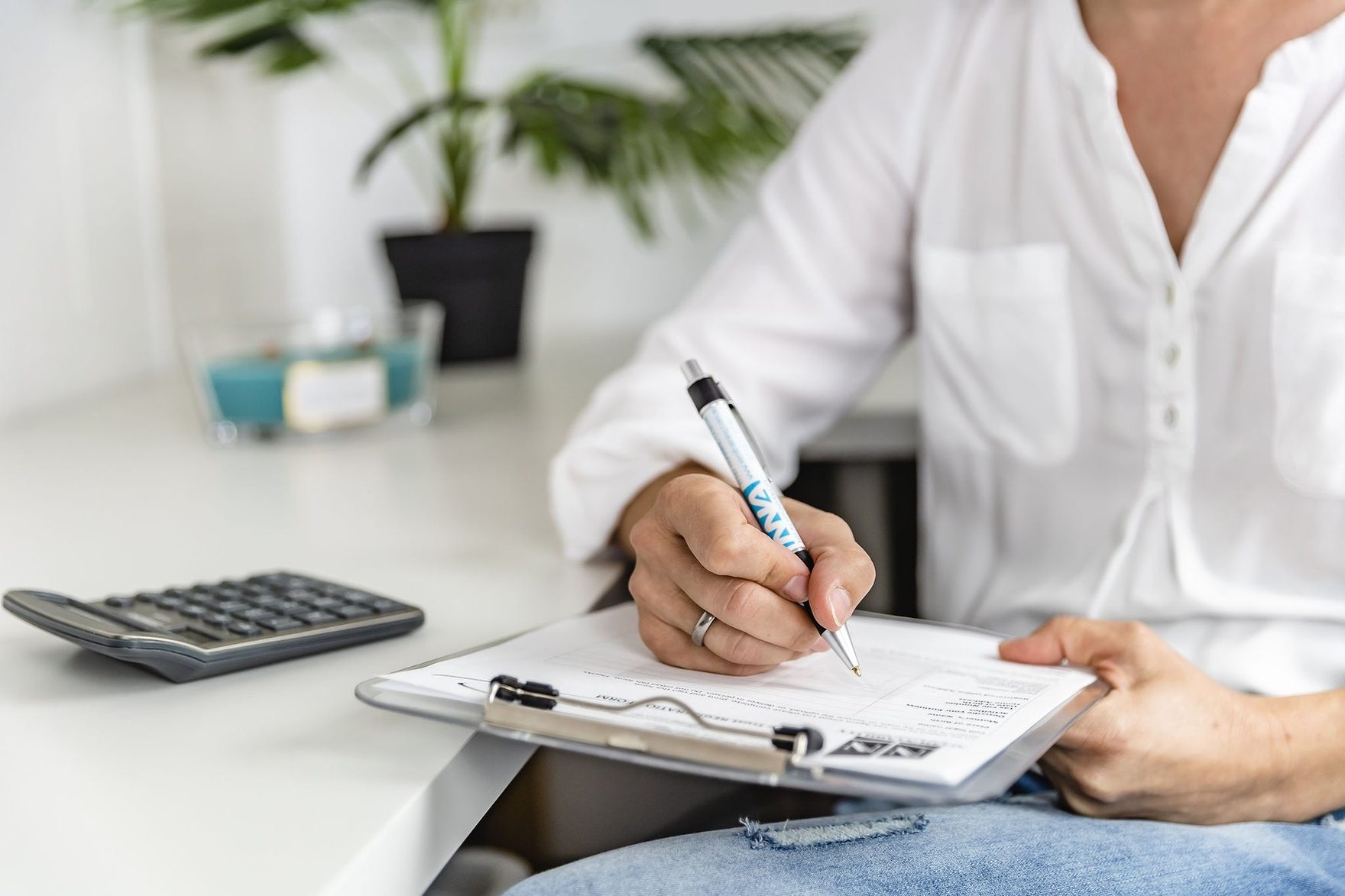 Hands writing on a clipboard form beside a calculator at a white desk