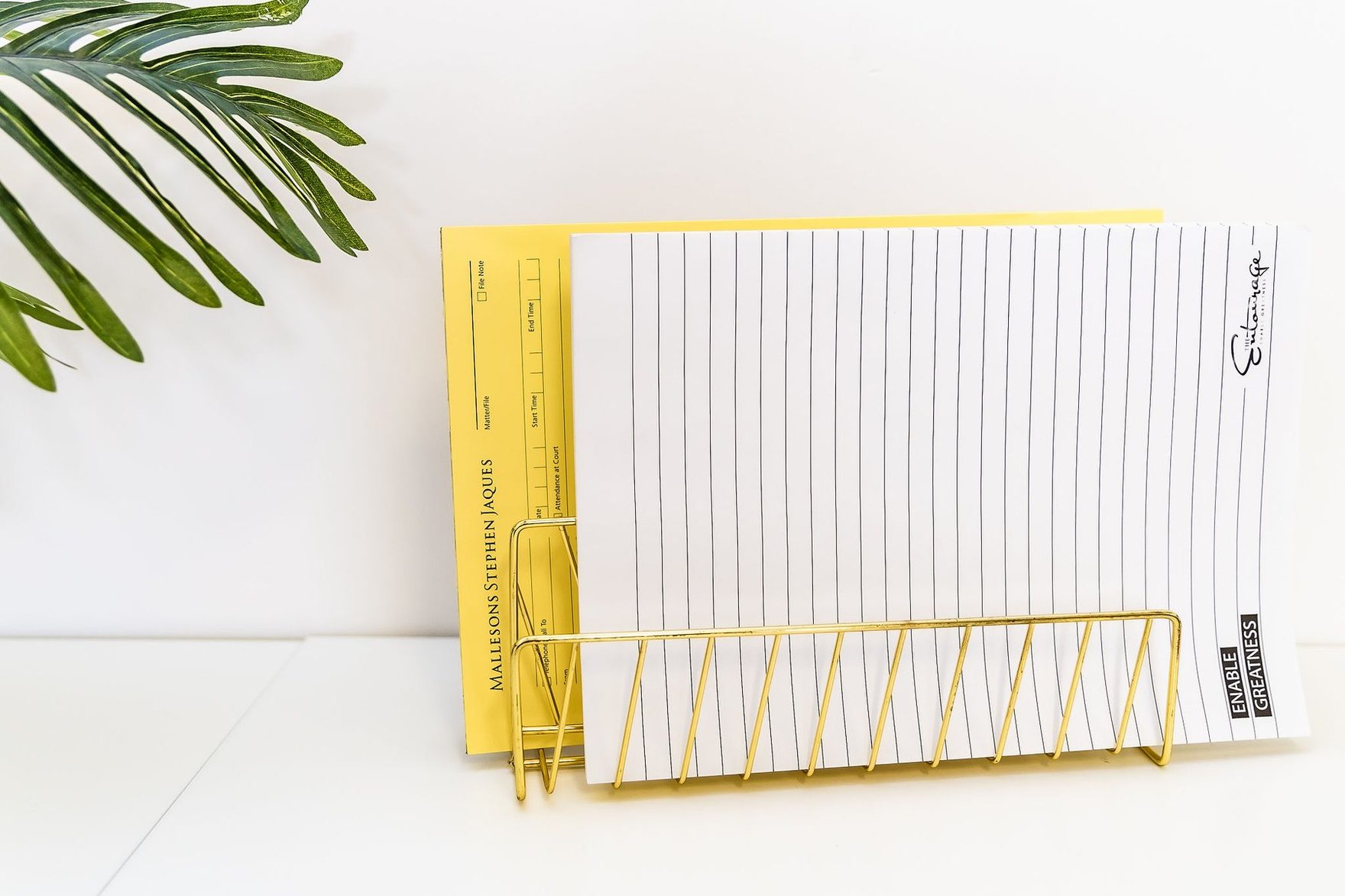 A gold wire file holder with notepads, beside a palm leaf