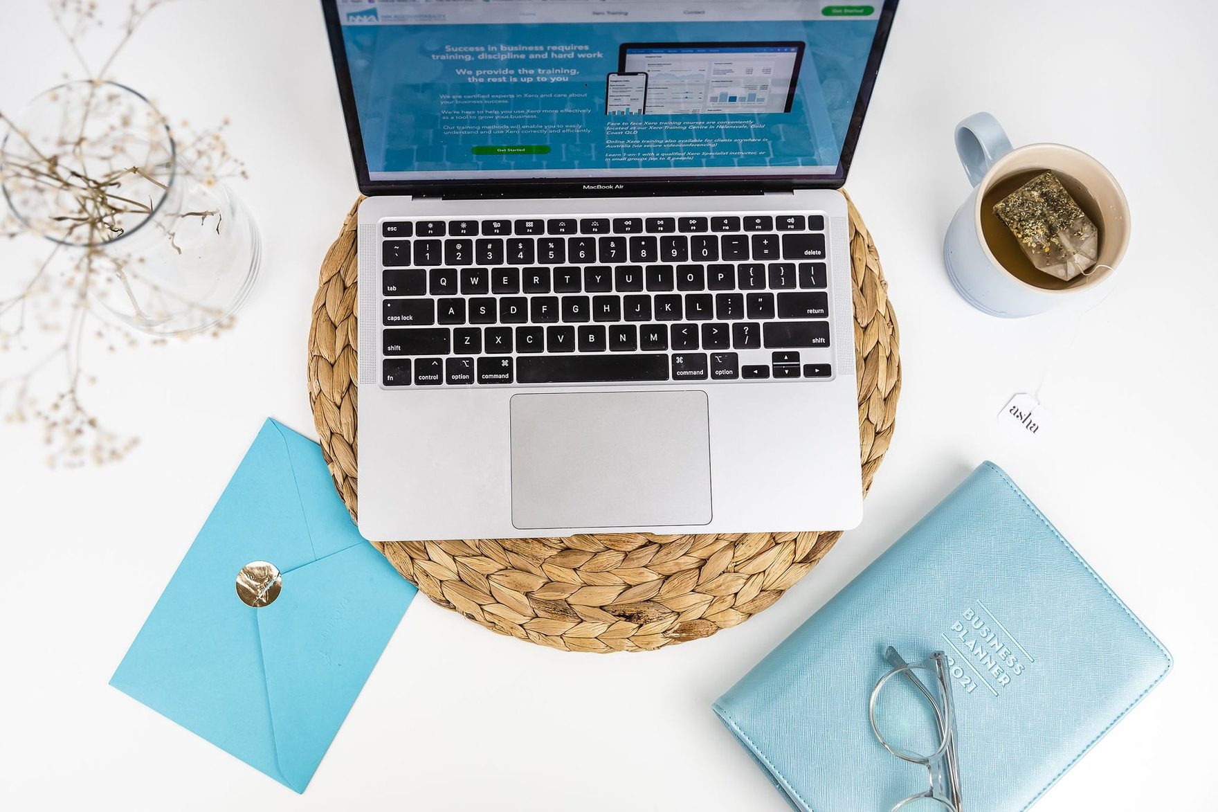 Overhead view of an open laptop on a woven mat, with a blue planner, an envelope and a cup of coffee